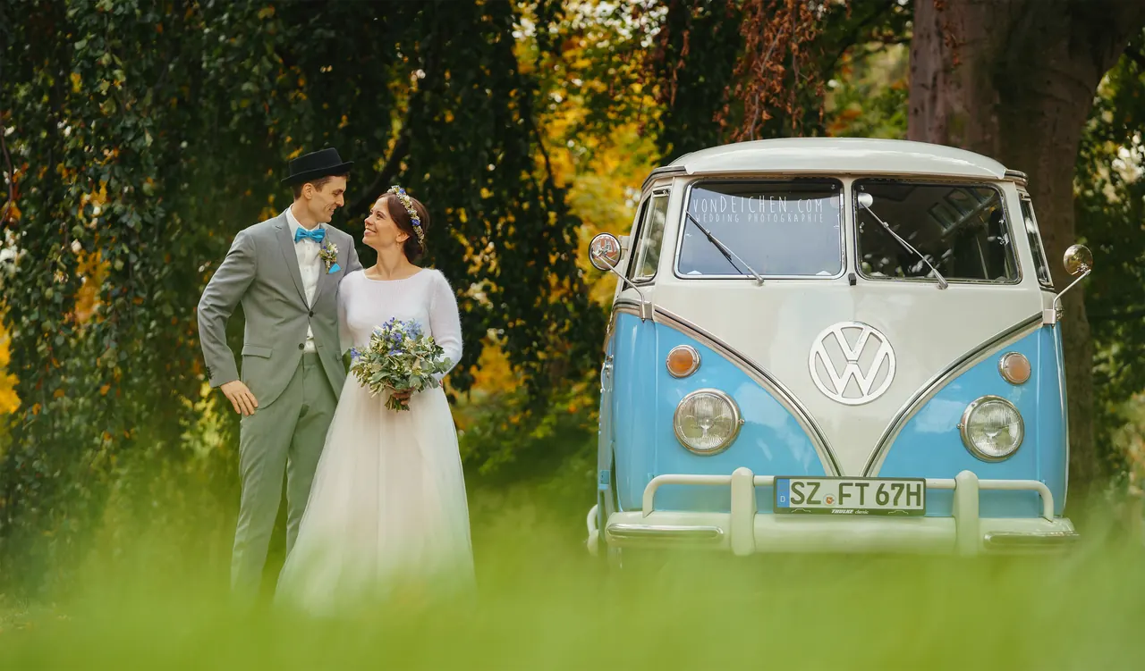 Brautpaar neben einem blauen VW-Bus im Grünen